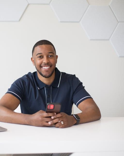 A smiling applicant at a desk with a laptop, holding his phone