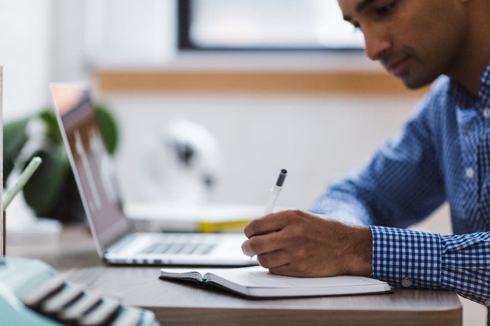 A man writing a checklist in a notebook beside his laptop